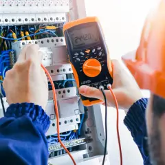 Elektryk w pomarańczowym kasku sprawdza napięcie multimetrem w skrzynce z przewodami.