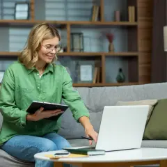Kobieta w okularach siedzi na kanapie, pracując na laptopie. To jej sposób na doładowanie energii online, planując kolejne zadania.