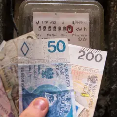 Dłoń trzyma banknoty obok licznika energii, symbolizując rosnące ceny energii elektrycznej.