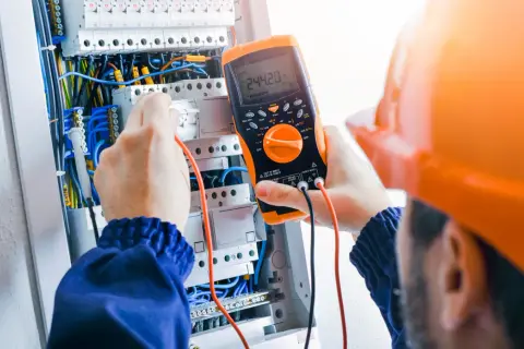 Elektryk w pomarańczowym kasku sprawdza napięcie multimetrem w skrzynce z przewodami.