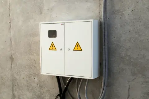 Biała skrzynka elektryczna z żółtymi symbolami ostrzegawczymi, gotowa do podłączenia prądu do domu.