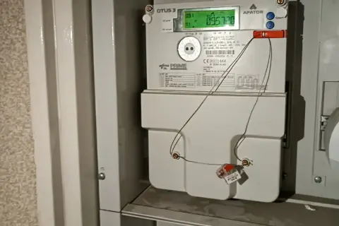 Cyfrowy licznik energii elektrycznej APATOR OTUS 3 z zielonym wyświetlaczem. Widać przewody podłączone do plomby, co może sugerować próby manipulacji, jak oszukać licznik prądu.