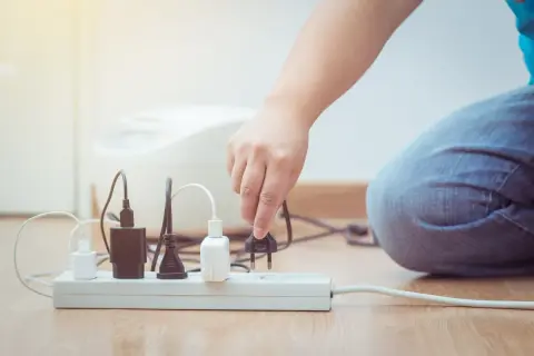 Ręka podłącza wtyczkę do listwy zasilającej, szukając ulgi na prąd. Wiele urządzeń już czeka na energię.