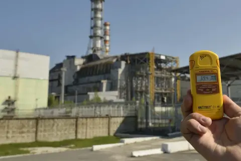 Żółty dozymetr pokazuje 3.94 µSv/h przed budynkami elektrowni w Czarnobylu, odpowiadając na pytanie, czy Czarnobyl dalej promieniuje.