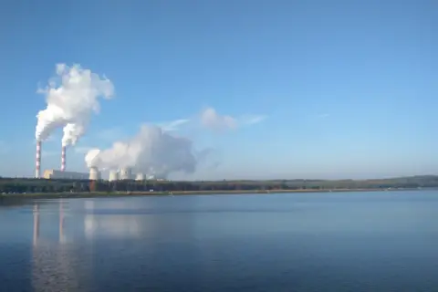 Dymiące kominy elektrowni w Bełchatowie, pokazujące, ile energii produkuje Bełchatów. W tle jezioro i las.