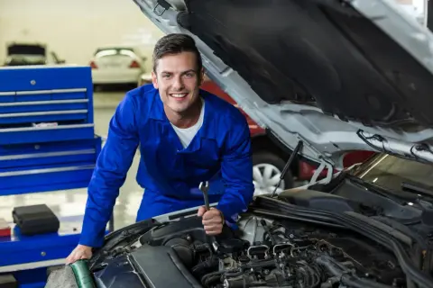 Uśmiechnięty mechanik w niebieskim kombinezonie z kluczem w dłoni, pracujący przy silniku. Dowiedz się, jak zostać elektrykiem samochodowym.