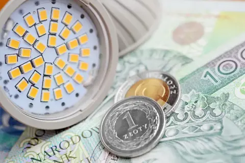 Żarówka LED i monety na banknocie 100 zł. Symbolizuje to wniosek o zamrożenie cen prądu dla gospodarstw domowych.