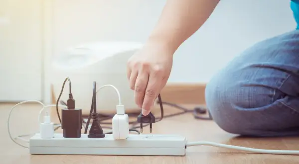 Ręka podłącza wtyczkę do listwy zasilającej, szukając ulgi na prąd. Wiele urządzeń już czeka na energię.