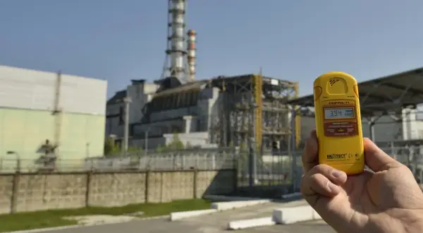 Żółty dozymetr pokazuje 3.94 µSv/h przed budynkami elektrowni w Czarnobylu, odpowiadając na pytanie, czy Czarnobyl dalej promieniuje.
