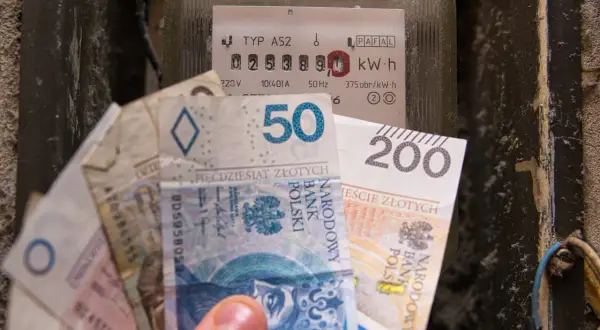 Dłoń trzyma banknoty obok licznika energii, symbolizując rosnące ceny energii elektrycznej.