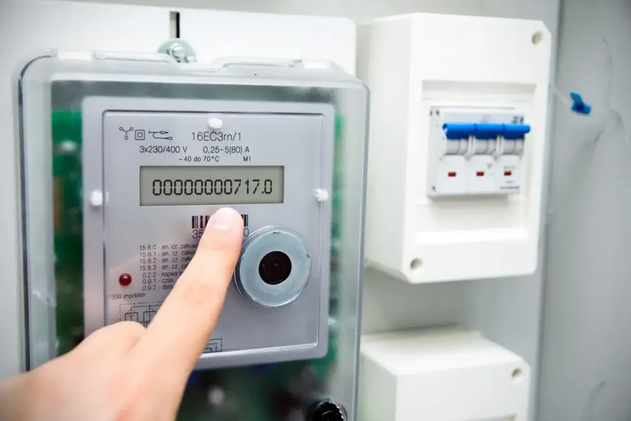 Palec wskazuje na odczyt licznika elektrycznego: 00000000717.0 kWh. Obok widoczny wyłącznik nadprądowy.