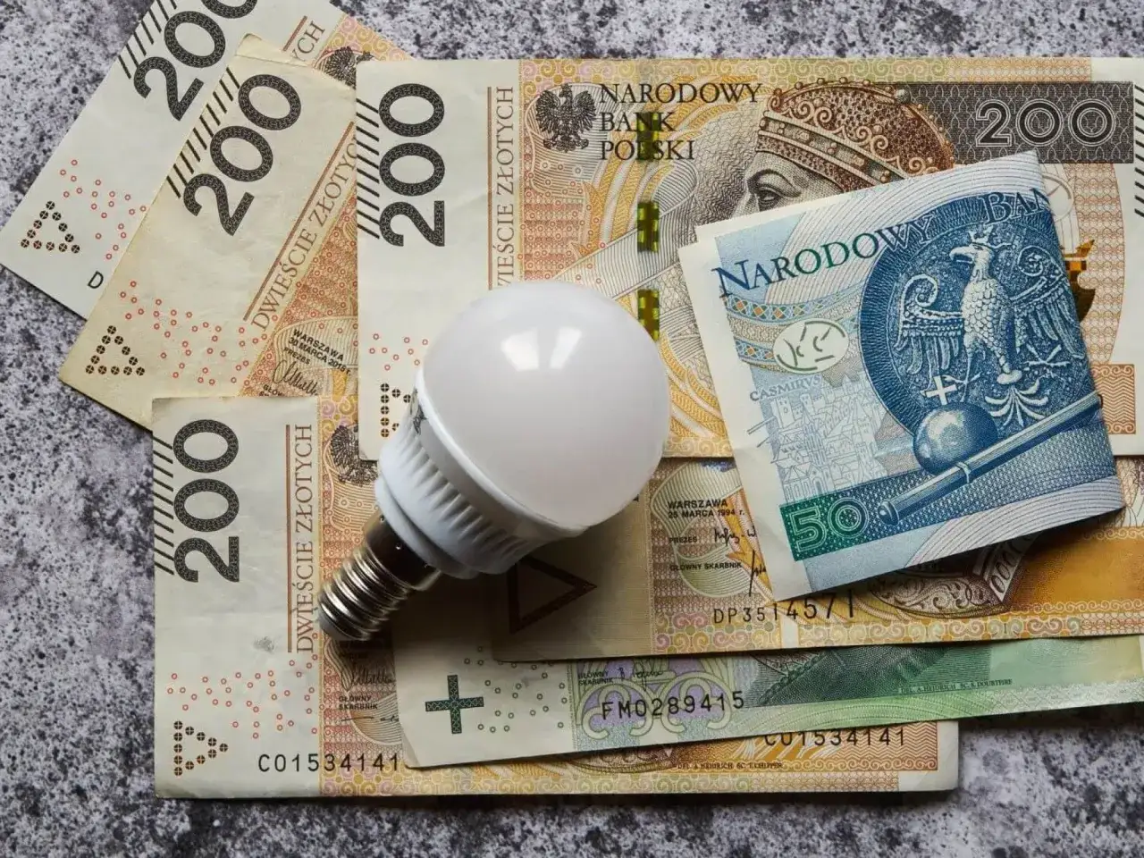 Biała żarówka LED leży na stosie polskich banknotów 200 zł i 50 zł. Symbolizuje obniżkę za prąd.