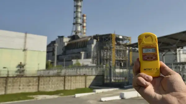 Żółty dozymetr pokazuje 3.94 µSv/h przed budynkami elektrowni w Czarnobylu, odpowiadając na pytanie, czy Czarnobyl dalej promieniuje.